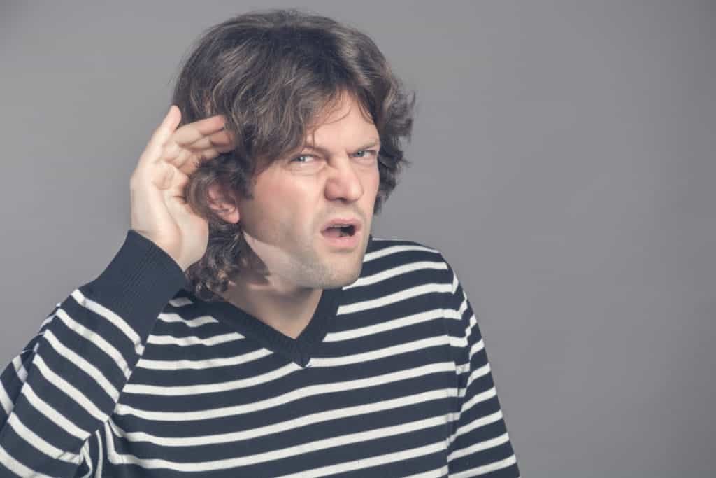 Unhappy hard of hearing man placing hand on ear asking someone to speak up or listening to bad news isolated on gray background. Negative emotion facial expression feelings. Angry male trying to hear